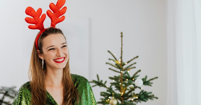Žena se smeje pored novogodišnje jelke i ima rajf na glavi u obliku rogova irvasa.