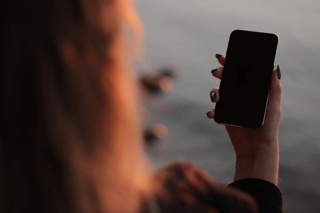 Ruka drži mobilni telefon sa crnim ekranom iznad vode pri zalasku sunca